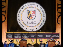 UMBC President and Provost standing on a staff behind podiums in their regalia with the UMBC seal on a backdrop behind them.