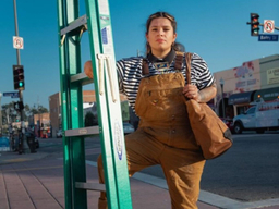 Johanna Toruño poses for an image on a busy street while leaning against a large ladder that appears to be against a building that is out of frame. She is wearing khaki overalls.