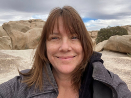 A woman with medium length brown hair and bangs smiles in front of a scenic backdrop of rocks, clouds, and sky.