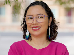 Yvette Martínez-Vu, a Chicana woman, is smiling. She is wearing a magenta-colored shirt and has dangly earrings.
