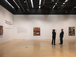 Two people stand in an art gallery looking at artworks hung on white walls.