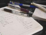 Notebook open to a page of class notes with a pile of different color pens overlapping and next to the notebook.