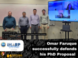 PhD committee standing with PhD Candidate after successful defense of proposal. 4 people in person, 2 people on TV Screen