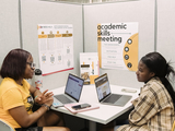 Two students sitting across the table from each other with their laptops out. They are in a cubicle and there are posters on the wall saying, "Academic Skills Meeting".